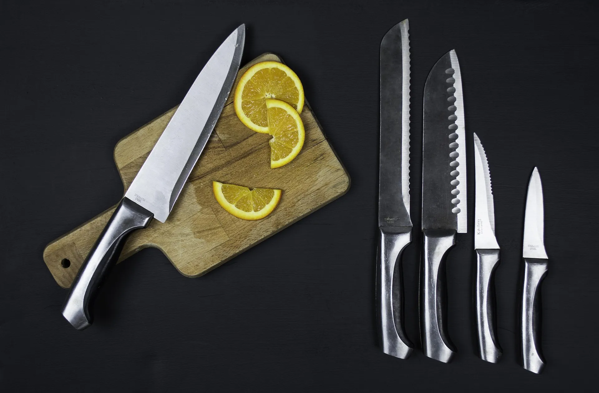 A chef's knife resting on a wooden cutting board beside orange slices, with four more kitchen knives laid out on a dark surface