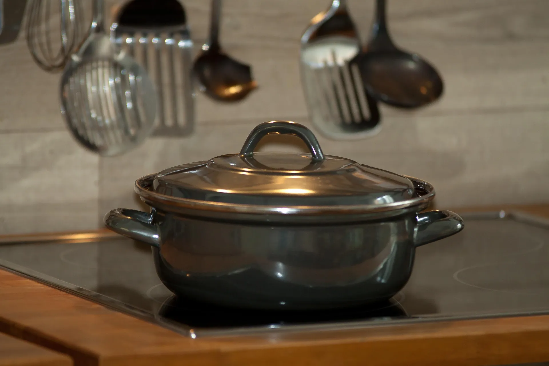 A dark enameled pot with a lid on a cooktop, with kitchen utensils hanging behind it