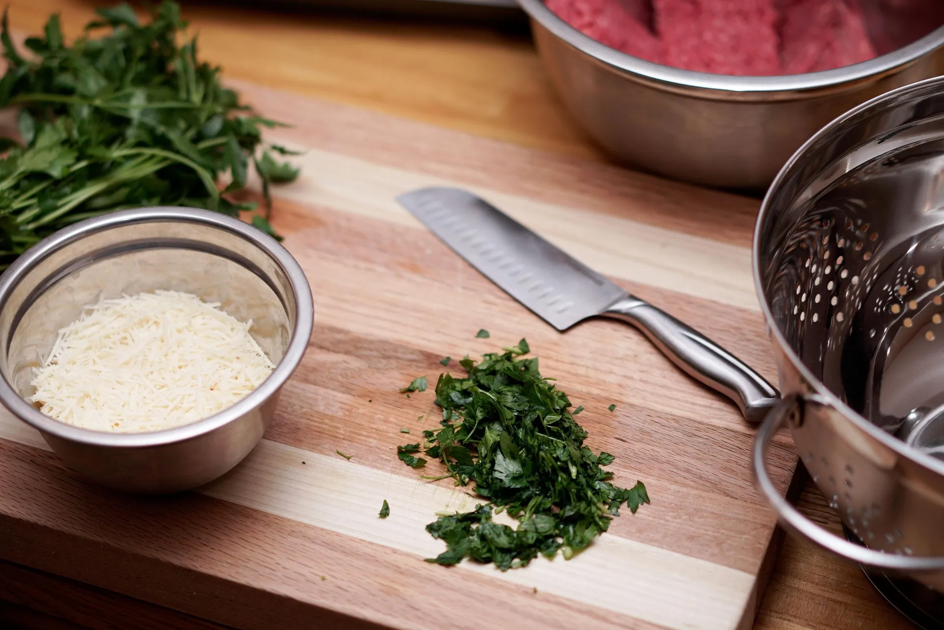 A wooden cutting board with chopped parsley, a bowl of grated cheese, and a santoku-style knife