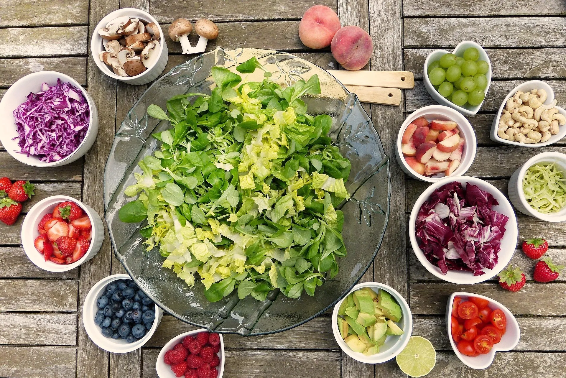 A large glass platter of salad greens surrounded by small white bowls of cut fruit and vegetables on a wooden table