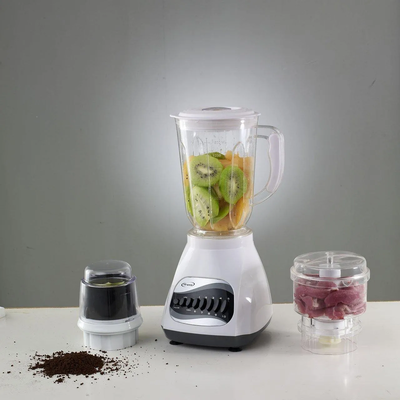 A white countertop blender holding sliced kiwi and melon, with a coffee grinder cup and a small chopper bowl of sliced meat beside it