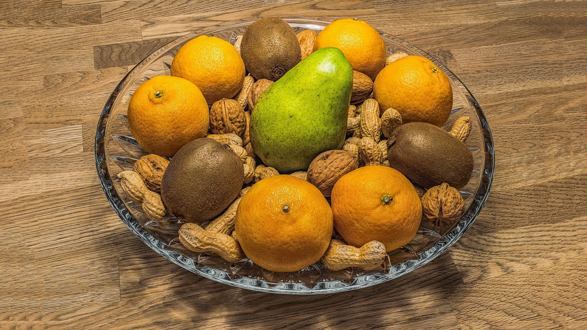A glass bowl holding oranges, kiwis, a pear, and nuts on a wooden table