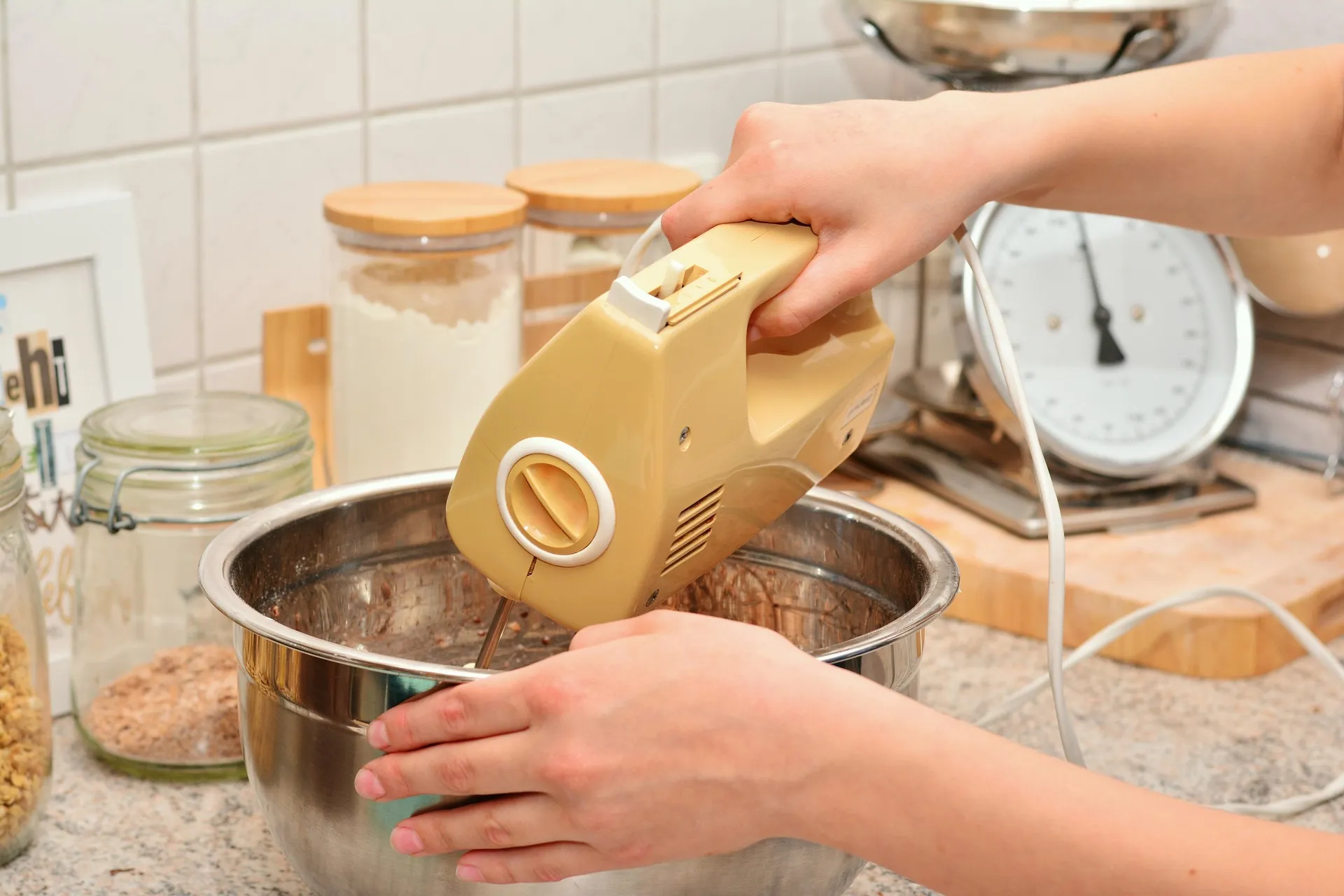 Hands holding a hand mixer over a stainless steel mixing bowl on a kitchen counter