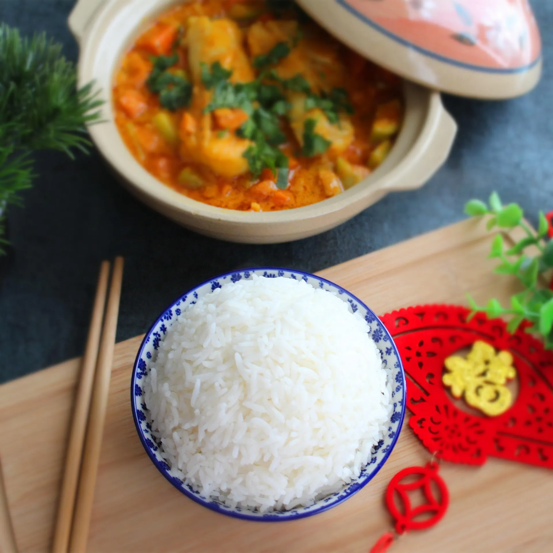 A blue-and-white bowl of cooked white rice beside a clay pot of vegetable curry on a wooden board