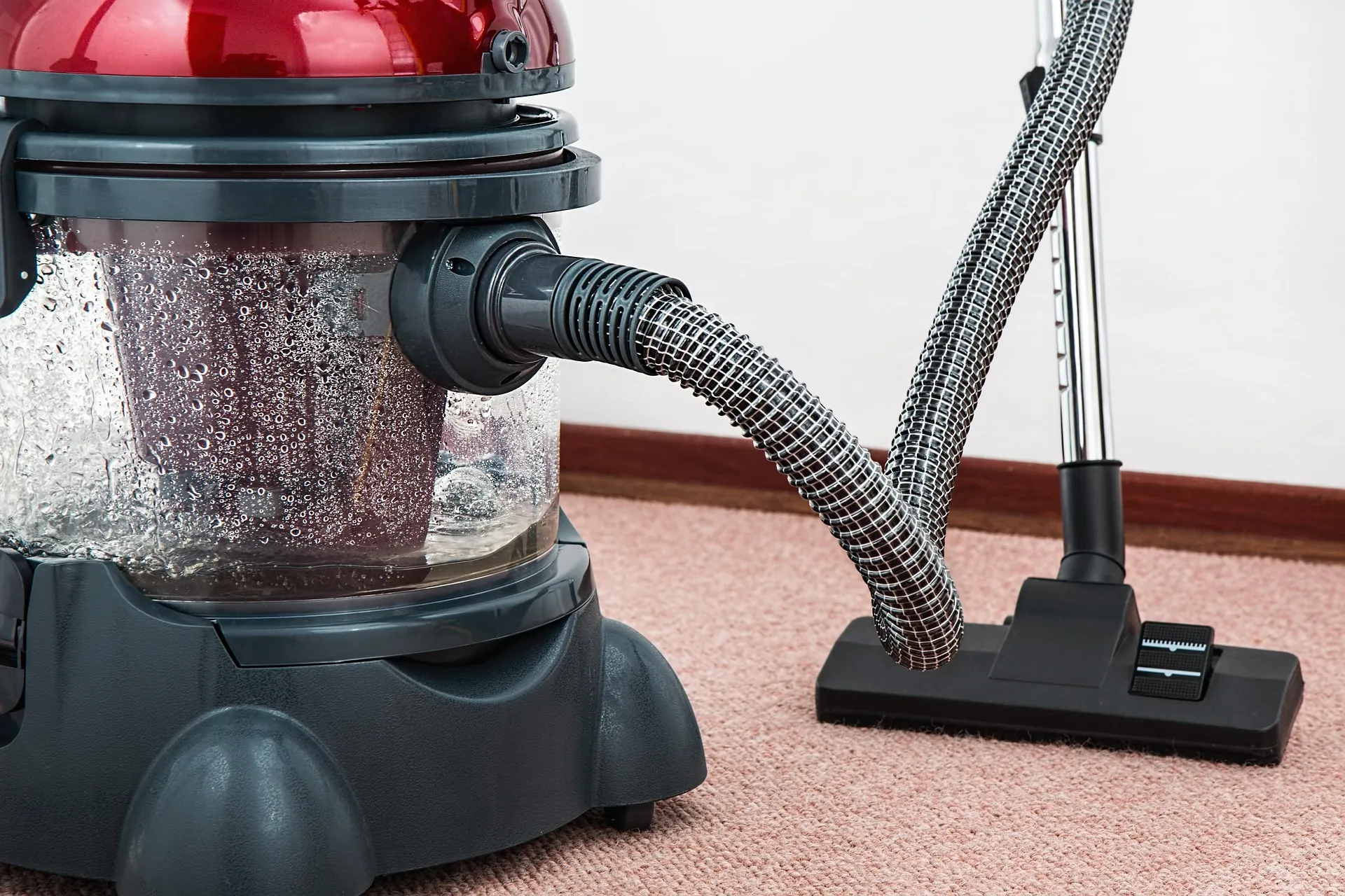 A red and gray canister vacuum cleaner with a transparent tank and ribbed hose on a pink carpet
